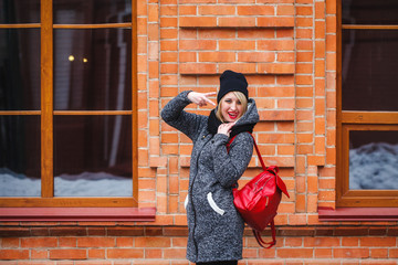 Young modern girl hipster shows a gesture with her hands and shows tongue. Portrait of a hipster girl on a red brick wall background.