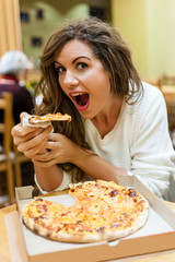 A young woman sitting in a restaurant, smiling,looking at the camera biting a slice of pizza.