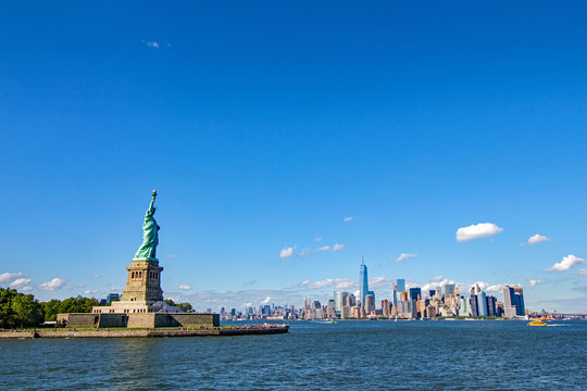 Statue of Liberty in foreground with unmistakeable New York’s Manhattan cityscape in background viewed across the Hudson and against a bright blue sky