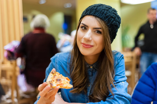 A young woman sitting in a restaurant, smiling,looking at the camera holding a slice of pizza.