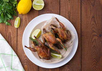 Appetizing baked quail with spices, herbs and lemon, lime in white dish on a wooden table Flat lay