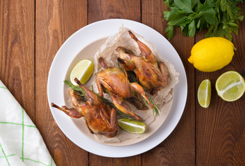 Appetizing baked quail with spices, herbs and lemon, lime in white dish on a wooden table Flat lay