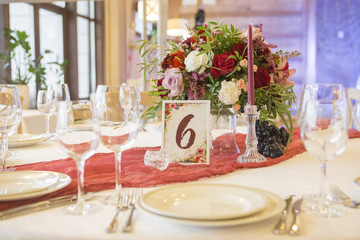 Set of glassess and plates on a wedding table surrounded by light garland and wine color flowers and tablecloth. Interior. Copy space