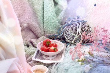 The girl is holding coffee. Fresh strawberries in a plate. On the sofa in the cozy living room. Accessories for hobby knitting. Spring morning. Bouquet of flowers. Card. Free space for text.