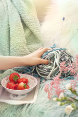 Girl holding fresh strawberries in a plate. On the sofa in the cozy living room. Accessories for hobby knitting. Spring morning. Bouquet of flowers. Card. Free space for text.