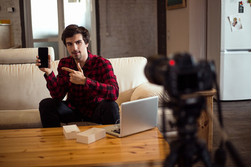 Fototapeta premium Young smart bearded man making a video for his own blog and advertising the smartphone on the white leather sofa at home.
