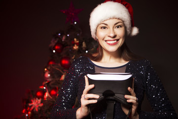 Cute smiling woman in Christmas hat and evening sexy dress presents VR glasses. New Year tree with red light on a background