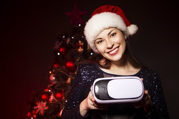 Cute smiling woman in Christmas hat and evening sexy dress presents VR glasses. New Year tree with red light on a background