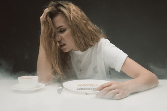 Depressed young girl feeling bad about smoking a lot. She is sitting with crooked face in front of plate full of cigarettes. Isolated on background