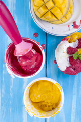 Fruit ice cream with mango and raspberries in a bucket pack. Spoon for ice cream. Blue background and flowers. Sweet dessert.