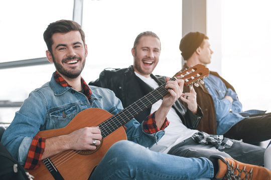 Laughing friends situating in room. Outgoing male performing melody with musical instrument. Friendship concept