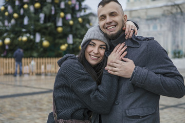 Gorgeous beautiful caucasian woman with a man on a walk in european city at winter. Couple wear casual grey outfit and hugging each other. Cold weather, christmas lights on a background