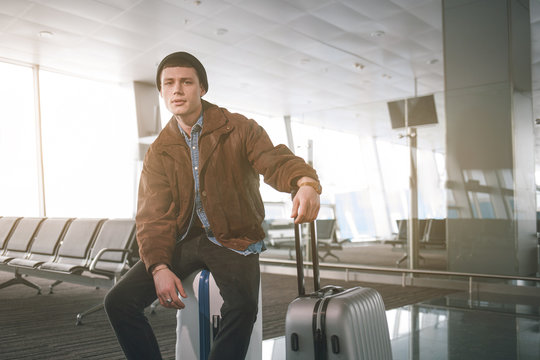 Portrait of weariful young man sitting on luggage while looking in camera in airport. Anticipation concept