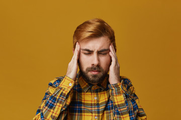 Portrait of young guy keep his eyes closed holds fingers on temples feels his head hurts very bad, wears yellow plaid shirt. Isolated over yellow background