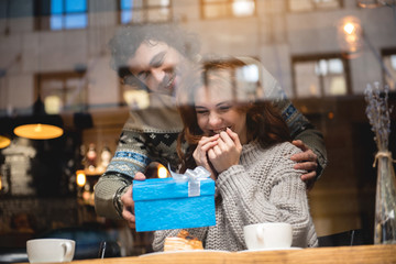 This is for you. Cheerful man is giving present to his wife in cafeteria. Woman is looking at blue wrapped box and smiling. View from glass window
