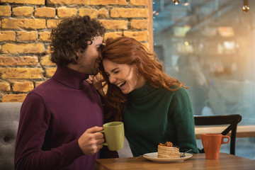 Joyful loving couple is entertaining in cafe together. They are drinking tea with cake and laughing