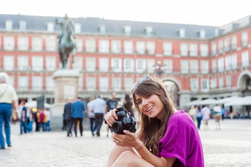 Young woman tourist holding a photo camera and taking picture in Plaza Mayor square, Madrid, Spain. Tourist attraction, statue of Felipe III in the background.