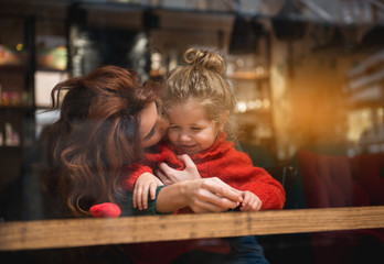 Cheerful woman is kissing her little daughter with love. She is cuddling the smiling girl while...