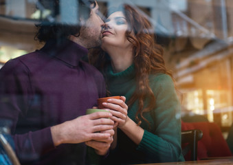 Glad young man and woman touching their noses with fondness. They are keeping cups of hot tea and smiling. View from the window