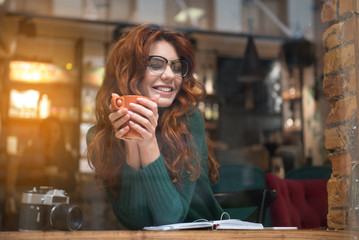 Portrait of happy young woman enjoying coffee in cozy cafeteria. She is keeping mug in both hands and laughing. Relaxation concept