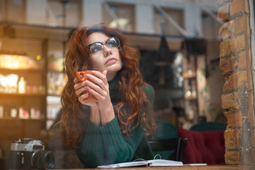 Dreamful red-haired girl drinking hot beverage in cafe. She is sitting at table and looking up pensively