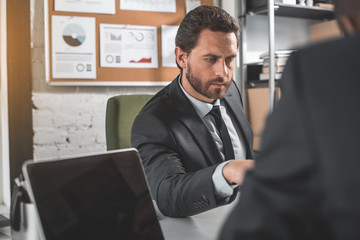 Fototapeta premium Business plan. Serious young bearded man in suit is sitting at table and expressing confidence while laboring with his colleague