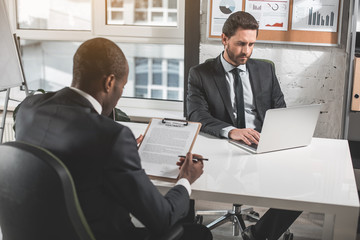 Important contract. Young bearded businessman is sitting at table with his colleague and looking at screen of laptop. Back view african man is reading document. Selective focus