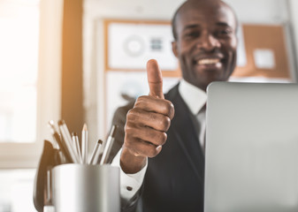 Its OK. Close-up of african businessman hand is giving thumbs-up to camera. Man is smiling on background while sitting at table in office. Focus on hand