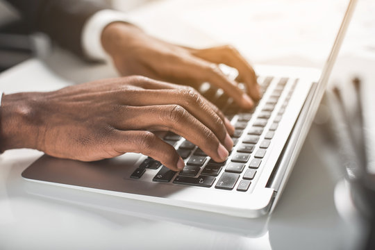 Modern device. Close up of hands of young african businessman is sitting at table and typing on laptop. Selective focus