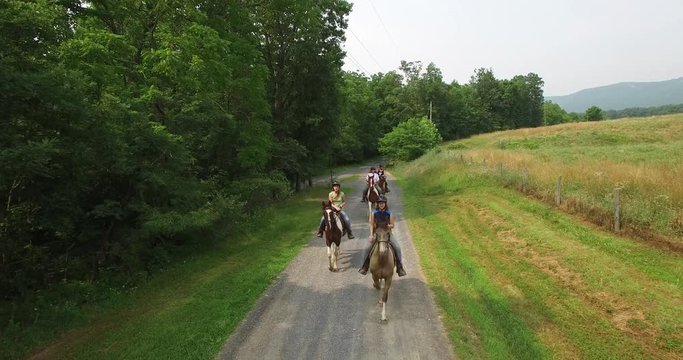 Aerial camera facing horseback riders coming towards camera and then flying above them on a dirt road lined with trees in summer.