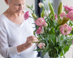 Older woman arranging pink lisianthus flowers with greenery in vase (selective focus)