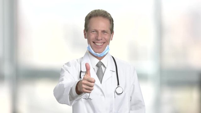 Happy male doctor gesturing thumb up. Cheerful mature doctor showing thumb up on blurred background. Symbol of success.