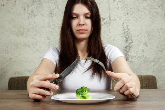 sad young brunette woman dealing with anorexia nervosa or bulimia having small green vegetable on plate. Dieting problems, eating disorder.