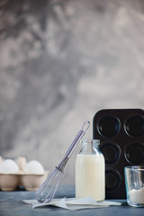 Cooking concept with baking tools and ingredients. Flour, a muffin tin, whisk, a bottle of milk and a carton of eggs on a concrete background. High key food photography with copy space.