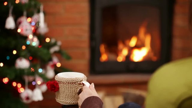 Woman sitting and relaxing by fireplace in a rocking chair holding a cup of hot drink at christmas time - camera dolly, closeup