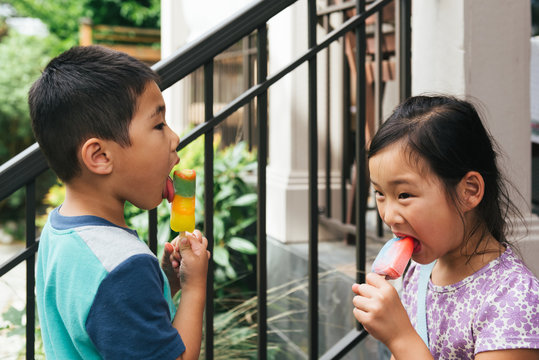 Asian american brother and sister eating popsicles facing each other on porch steps