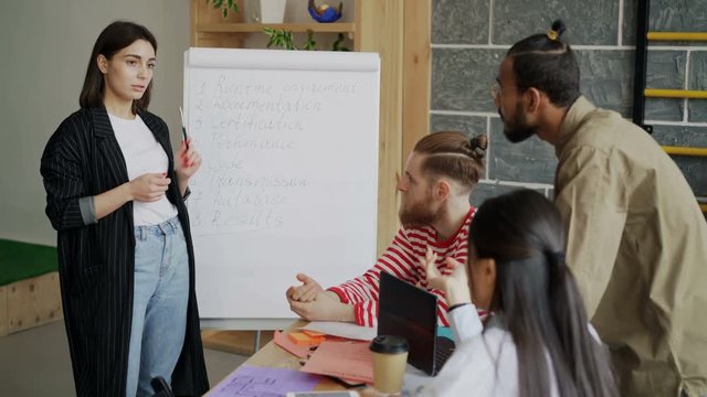 Cheerful female entrepreneur explaining start-up software development brief on flipchart to multi ethnic colleagues in modern loft office indoors