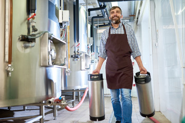 Portrait shot of smiling middle-aged worker wearing apron carrying heavy beer kegs while walking along corridor of modern brewery