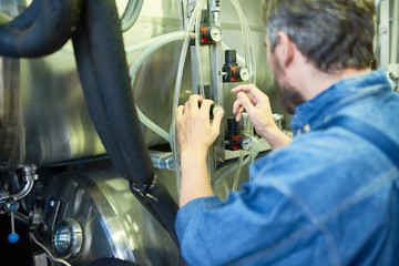 Back view of bearded brewer wearing denim shirt adjusting pressure in cylindrical beer maturation tank while working at modern brewing plant