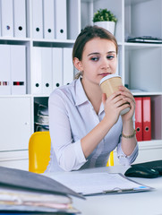 Tired and exhausted woman looks at the mountain of documents holding a paper glass with coffee in hands.