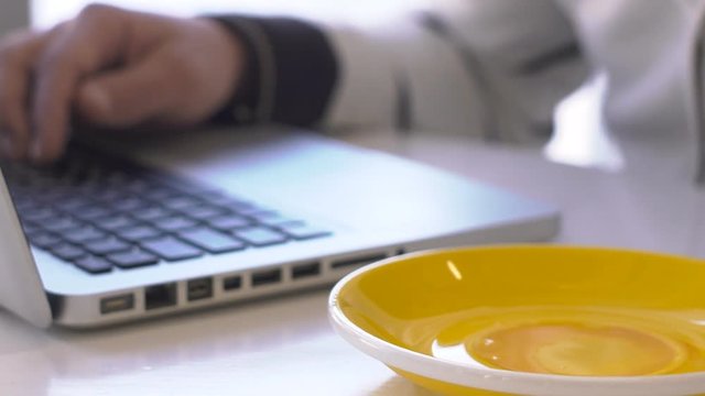 Close-up of man working on laptop and drinking tea from mug with saucer. While working and searching for information on the Internet, a male person takes a sip of a warm drink from jorum and an orange