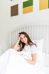 Positive young woman with pillow sitting in bed and smiling to camera at home