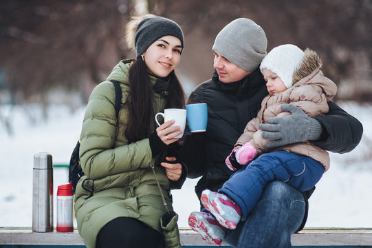 Young parents with their little daughter drinks hot tea from a thermos in the winter. Winter walk. Happy young family on snow background.