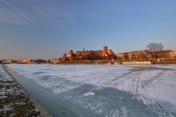 Piękny zimowy pejzaż zamku na Wawelu w Krakowie z zamarzniętą rzeką Wisłą na pierwszym planie, światło zachodzącego słońca, na lodzie na rzece ślady sanek i butów - obrazy, fototapety, plakaty
