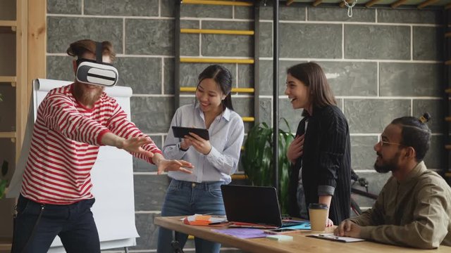 Mult ethnic team testing VR game simulator at modern office. Bearded hipster man using virtual reality headset feels fun while asian girl use gaming tablet