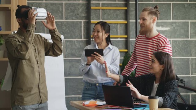 Mult ethnic team testing VR game simulator at modern office. African american man trying play virtual reality headset while asian girl using digital tablet