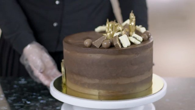 Female confectioner is observing delicious chocolate cake at the kitchen, close up. Woman in black uniforn and white gloves is turning around the plate. Signs Happy birthday and 19 are made with