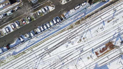Perpendicular aerial view of the tracks of a subway station. At the side of the tracks there is a city street with parked cars. Everything is covered by snow on this winter day.