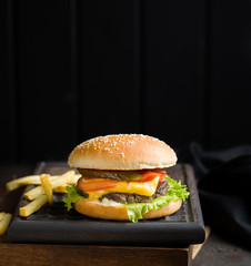 Delicious fresh tasty burger with beef, tomato, cheese, lettuce and french fries served on a wooden cutting board on dark background. Street fast food