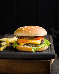 Delicious fresh tasty burger with beef, tomato, cheese, lettuce and french fries served on a wooden cutting board on dark background. Street fast food